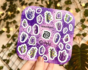 Op de afbeelding: Een vierkant stickervel met illustraties van amethistkristallen in paarse tinten, met groene en gele accenten. Het woord "amethyst" staat op het vel, samen met een spiraalontwerp en de handtekening van de kunstenaar.
