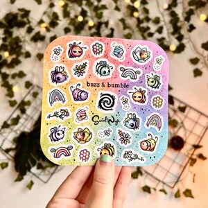 May include: A sheet of colorful bee-themed stickers with a rainbow gradient background. The stickers feature various bees, flowers, honeycombs, and rainbows. The text "buzz & bumble" is printed on the sheet.