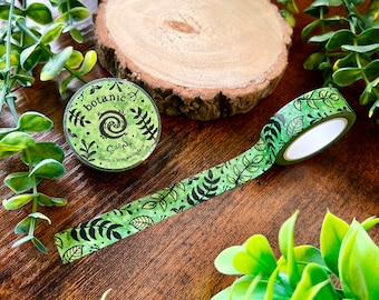 May include: A roll of green washi tape with a black and white botanical pattern. The tape is 15mm wide and 10m long. The tape is labeled "botanic" and "Crafty" with a small snail graphic.