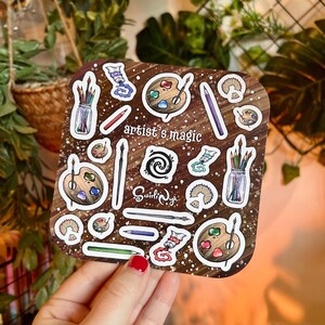 May include: A sticker sheet with a brown background and white speckles. The sheet features illustrations of art supplies, including paint palettes, paint tubes, pencils, paintbrushes, and the text "artist's magic".