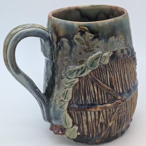 May include: A ceramic mug featuring a glossy blue and brown glaze with a textured relief design of a wooden door surrounded by sculpted green leaves. The mug has a curved handle and a smooth interior rim, showcasing a blend of earthy tones.