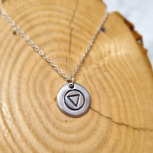 May include: A silver necklace with a circular pendant. The pendant has a triangle inside a circle. The chain is delicate. The necklace is displayed on a wooden surface.