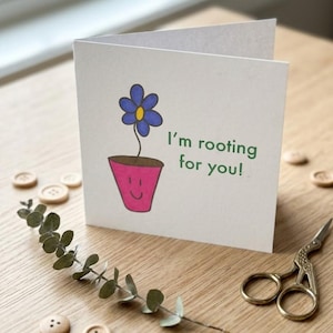 May include: A white greeting card featuring a blue flower in a pink pot illustration. The card reads "I'm rooting for you!" in green text. The card is on a wooden surface with decorative buttons, a sprig of greenery, and gold scissors.