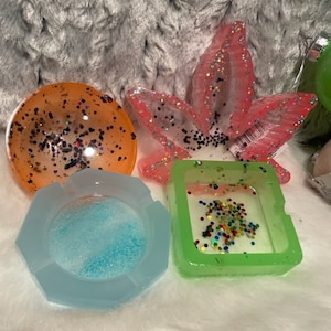 Könnte beinhalten: Vier Aschenbecher aus Harz mit Glitter, der in das Harz eingebettet ist. Einer ist ein runder orangefarbener Aschenbecher, einer ist ein quadratischer grüner Aschenbecher, einer ist ein hellblauer Aschenbecher mit sechseckiger Form und einer ist ein pinkfarbener Aschenbecher in Blattform.