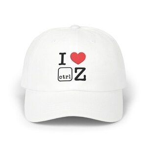 Op de afbeelding: Een witte baseballpet met een geborduurde afbeelding op de voorzijde. Het ontwerp toont de letter I, een rood hartsymbool, een vierkant icoon met de tekst ctrl en de letter Z, wat de sneltoets voor ongedaan maken weergeeft.