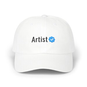 以下が含まれることがあります： 黒い文字でArtistと書かれ、その後に青い認証バッジのアイコンが付いた白い野球帽。この帽子は構造化されたクラウン、カーブしたつば、中央上部にボタンを備えており、シンプルで清潔感のあるデザインに仕上がっています。
