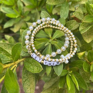 May include: Three beaded bracelets with white faux pearls and gold accents. One bracelet has the phrase "LET THEM" spelled out in white letter beads. The bracelets are arranged on a bed of green leaves.
