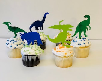 Op de afbeelding: Zes cupcakes versierd met witte frosting en kleurrijke sprinkles. Elke cupcake heeft een andere dinosaurus silhouet topper in blauw, groen en oranje.