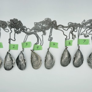 May include: Eight silver-toned metal mussel shell pendants on matching link chains. Each pendant is shaped like a textured bivalve shell and features a small green paper tag numbered one through eight attached to the chain near the top.