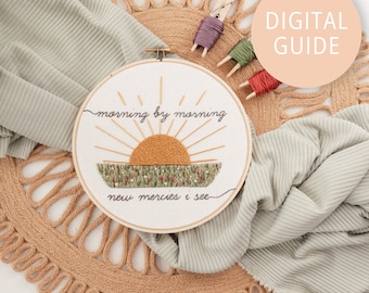 May include: A white embroidery hoop with a sunrise design and the text "morning by morning new mercies I see". The hoop is on a brown woven mat with a light green fabric draped over it. There are three embroidery floss skeins and a clothespin on the mat.