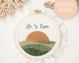 May include: Embroidered hoop art with a green and brown landscape, a setting sun, and the text "He Is Risen".