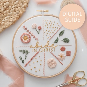 May include: A white embroidery hoop with a floral design and the text "abide in Christ". The hoop is surrounded by a light pink fabric and a woven straw mat.