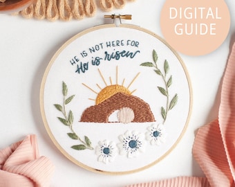 May include: Embroidered hoop art with a white background, featuring a brown tomb with a white stone rolled away, a yellow sun rising above the tomb, green leaves, and white flowers. The text "He is not here for He is risen" is embroidered in blue thread.