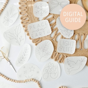 Peut inclure: Plusieurs patchs en tissu blanc avec des motifs brodés en gris, dont un flocon de neige, des feuilles, une colombe et les mots « Peace on Earth » et « Light & Life ». Une étiquette « Digital Guide » est visible. Les patchs sont disposés sur une surface texturée et beige.