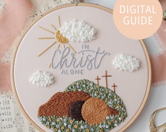 May include: Embroidered hoop art with a pink background featuring a sun, clouds, and the text "In Christ Alone". The embroidery also depicts a brown hill with three crosses and a stone tomb.