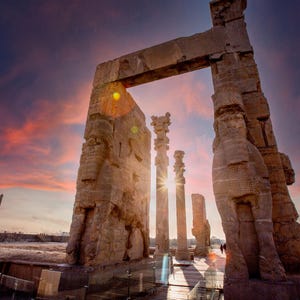 May include: A large stone archway with several pillars, all carved with intricate details. The archway is set against a vibrant pink and orange sunset sky.