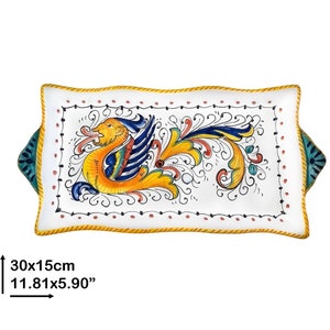 Könnte beinhalten: Ein weißes Keramik-Serviertablett mit einem handbemalten Design in Gelb, Blau und Rot einer mythischen Kreatur mit Flügeln. Das Tablett hat einen gewellten Rand und zwei Griffe. Das Tablett misst 30x15cm.