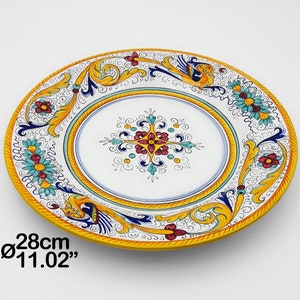 May include: A large, round, white ceramic plate with a yellow rim. The plate is decorated with a colourful floral pattern and a central cross design. The plate measures 28cm in diameter.