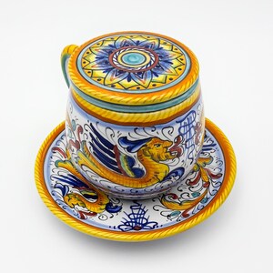 May include: A ceramic mug with a lid and a matching saucer. The mug and saucer are decorated with a colorful floral design and a winged dragon.
