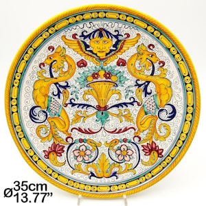 Op de afbeelding: Een grote, ronde, gele en witte keramische plaat met een decoratief ontwerp met twee gestileerde draken, een centraal bloemenmotief en een gezicht in een kroon. De plaat heeft een diameter van 35 cm.
