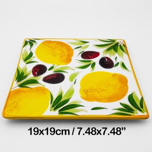 Puede incluir: Un plato cuadrado de servir de cerámica blanca con un borde amarillo. El plato presenta un diseño pintado de limones y aceitunas sobre un fondo blanco. El plato mide 19 cm por 19 cm.