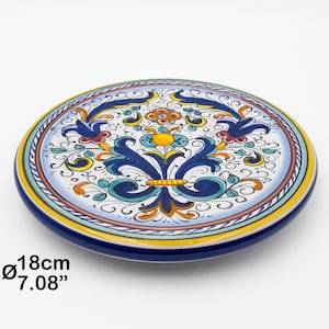 May include: A round ceramic plate featuring a hand-painted floral pattern in blue, yellow, orange, and green on a white background. The plate has a diameter of 7.08 inches. The design includes symmetrical scrollwork and central flower motifs.