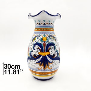 May include: A white ceramic vase with a scalloped rim and a blue, yellow, and orange floral design. The vase is 11.81 inches tall.