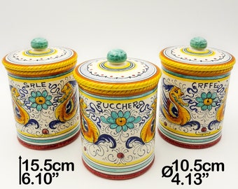Op de afbeelding: Drie cilindervormige keramische voorraadpotten met deksel, voorzien van handgeschilderde gele, blauwe en rode bloem- en fabeldiermotieven. De potten zijn 15,5 cm hoog met een diameter van 10,5 cm. Tekst op de potten bevat 'SALE', 'ZUCCHERO' en 'CAFFE'.