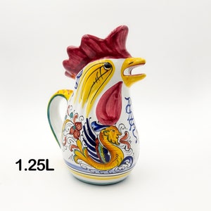 Puede incluir: Una jarra de cerámica blanca con forma de gallo y una capacidad de 1,25L. La jarra presenta una cresta roja, un pico amarillo y motivos florales y de criaturas míticas pintados a mano en azul, amarillo y rojo por todo el cuerpo.