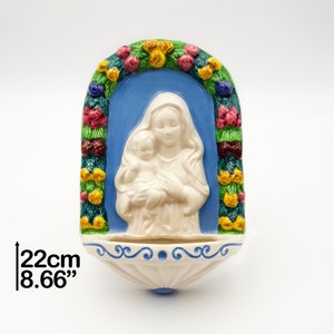 May include: A ceramic wall-mounted holy water font featuring a white relief of a figure holding a child against a blue background. The arch is decorated with colorful fruit and leaves. The base has blue scroll details and measures 22 cm or 8.66 inches.