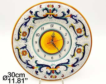 May include: A round wall clock with a 11.81 inch diameter, featuring a white face with black numerals and a central yellow circle. The border is decorated with blue, orange, and yellow floral scrollwork. It has black hour and minute hands.