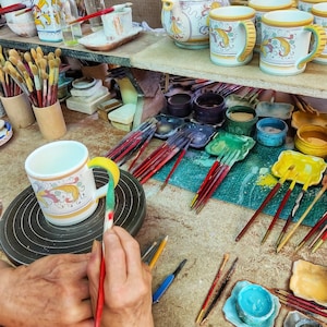 May include: A person paints a white ceramic mug with colorful floral designs. The mug is on a pottery wheel. There are paintbrushes, paint jars, and other ceramic pieces in the background.