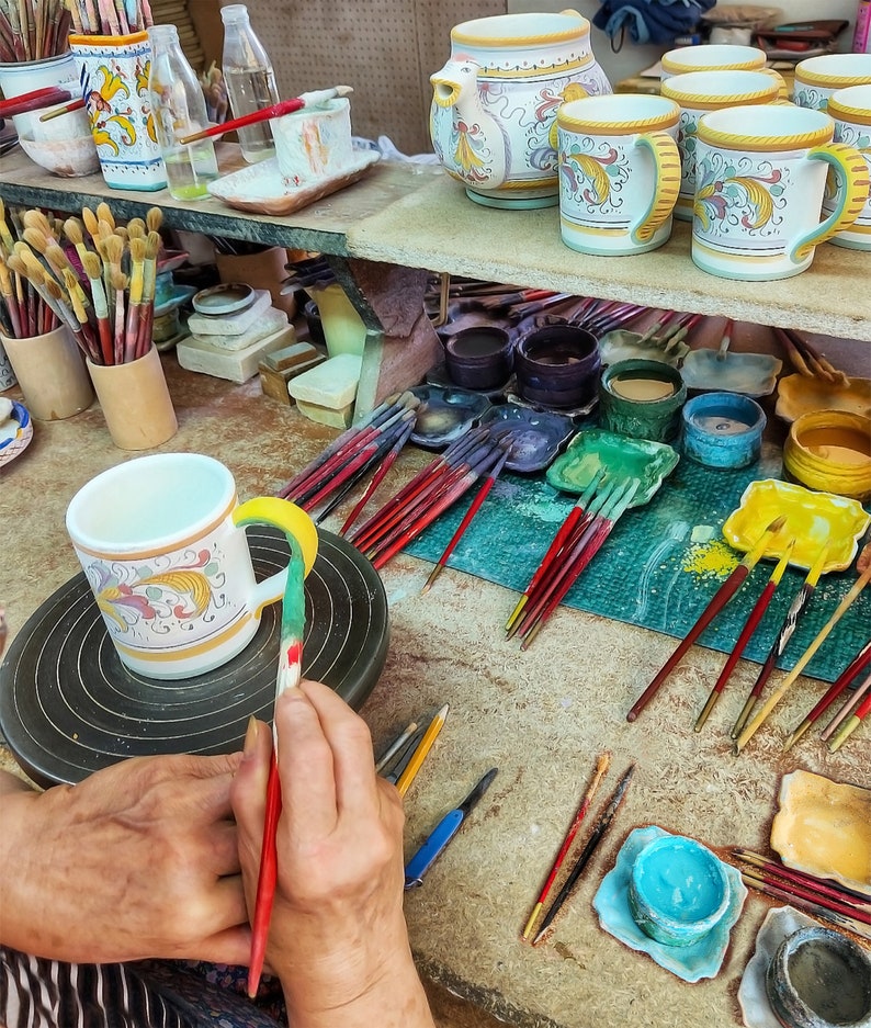 May include: A person paints a white ceramic mug with colorful floral designs. The mug is on a pottery wheel. There are paintbrushes, paint jars, and other ceramic pieces in the background.