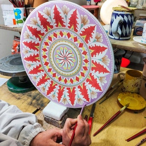 May include: A person paints a large, round ceramic plate with a colorful mandala design. The plate is decorated with red, yellow, green, and blue paint. The person is using a red paintbrush to add details to the design.