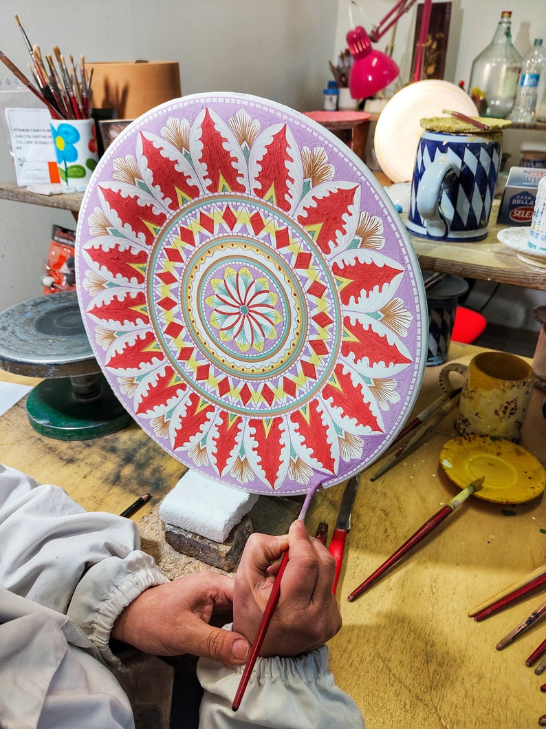 May include: A person paints a large, round ceramic plate with a colorful mandala design. The plate is decorated with red, yellow, green, and blue paint. The person is using a red paintbrush to add details to the design.