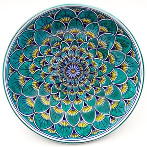 May include: A round ceramic plate with a blue, green, and yellow floral design. The design features a central flower with a blue center and yellow petals, surrounded by concentric rings of smaller flowers.
