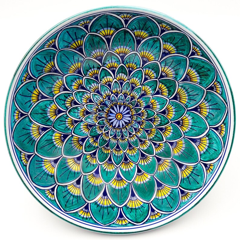 May include: A round ceramic plate with a blue, green, and yellow floral design. The design features a central flower with a blue center and yellow petals, surrounded by concentric rings of smaller flowers.
