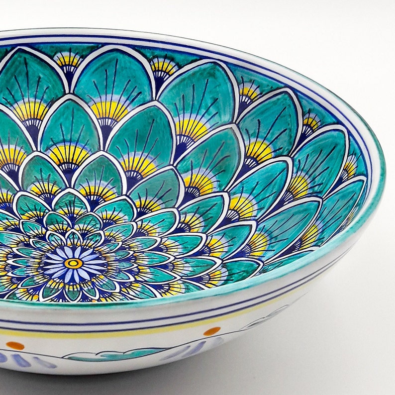May include: A large, white ceramic bowl with a blue, green, and yellow geometric pattern. The pattern features a repeating design of overlapping, fan-shaped elements.