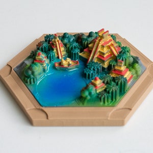 May include: A 3D printed miniature scene of a jungle with two golden pyramids, a blue lake, and green trees. The scene is framed in a hexagonal wooden frame.