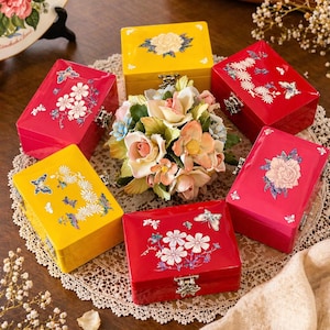 Può includere: Una collezione di scatole decorative in legno di vari colori, tra cui rosso e giallo, decorate con motivi floreali e farfalle. Ogni scatola ha un coperchio incernierato e una chiusura in metallo. Al centro c'è una composizione floreale.