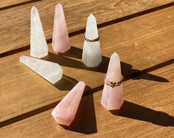 May include: A collection of crystal ring holders in clear and rose quartz. Two rings are displayed on the holders. The holders are arranged on a wooden surface, with sunlight casting shadows. The ring holders are faceted and cone-shaped.