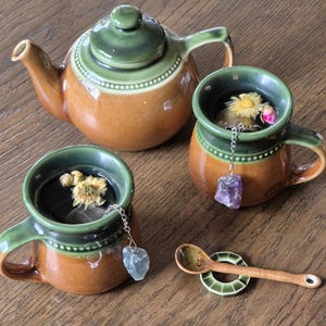 Puede incluir: Una tetera marrón y verde con tapa verde, dos tazas de té marrón y verde con infusores de té, una cuchara de madera, un platillo verde y dos piedras de cristal.