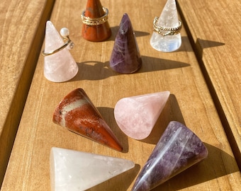 May include: A collection of seven polished stone ring cones in various colors including light pink, deep red, purple, and clear white. Three of the cones are displayed with gold-toned metal rings featuring pearls and textured bands.