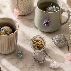 May include: Three mugs of tea with silver tea infusers and gemstone charms. The infusers are spherical and open to reveal loose tea. The mugs are beige and grey, and a small bowl of loose tea is also present.