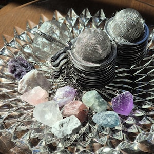 May include: A collection of colourful crystals and a set of silver tea infusers on a clear glass plate. The crystals are various shades of pink, purple, green, and white.