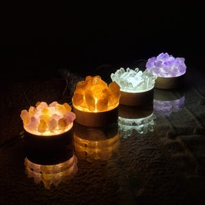 May include: Four crystal salt lamps with different coloured lights. The lamps are arranged in a row on a dark surface. The lamps are lit and casting a warm glow.