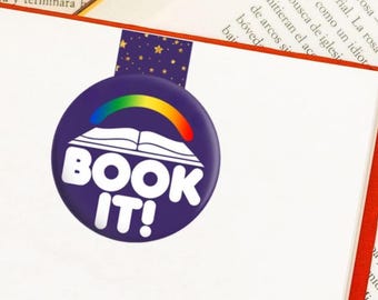 May include: A circular purple bookmark featuring a white open book graphic topped with a colorful rainbow arch and the text BOOK IT! in bold white letters. The bookmark has a rectangular tab at the top decorated with small gold stars.