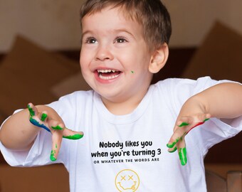 May include: A young boy wearing a white t-shirt with green paint on his hands. The shirt has the text "Nobody likes you when you're turning 3 OR WHATEVER THE WORDS ARE" and a smiley face with two X's for eyes.
