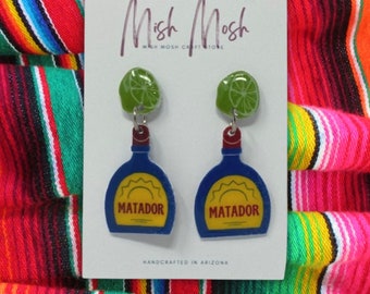 May include: A pair of handmade earrings featuring a blue bottle with a yellow sun and the word "MATADOR" on it, topped with a green lime slice. The earrings are on a white card with the text "Mish Mosh" and "HANDCRAFTED IN ARIZONA".