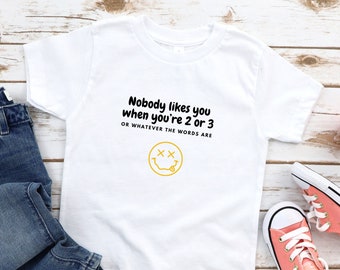 May include: White t-shirt with a yellow smiley face and the text "Nobody likes you when you're 2 or 3 OR WHATEVER THE WORDS ARE" in black.